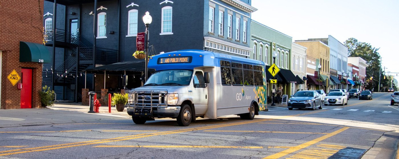A GoApex Route 1 bus with a blue roof and silver body on North Salem Street in downtown Apex, historic storefronts and a lamp post behind it