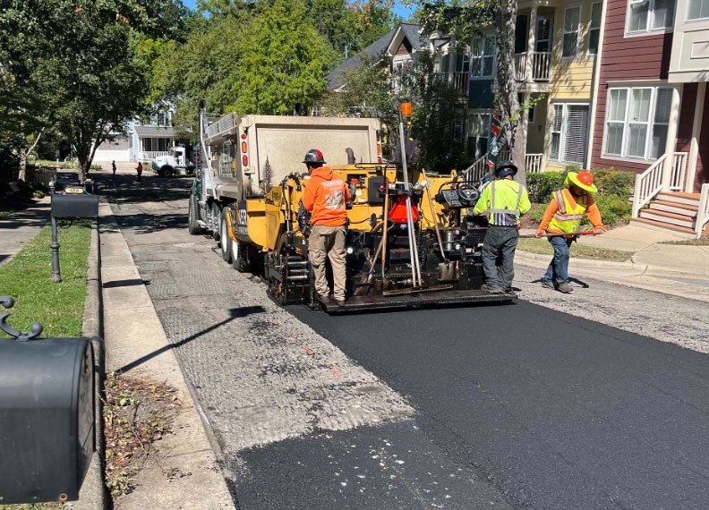 A paving crew in hi-vis vests runs an asphalt paver and dump truck, laying fresh black asphalt over a milled residential street lined with townhomes in Green at Scotts Mill
